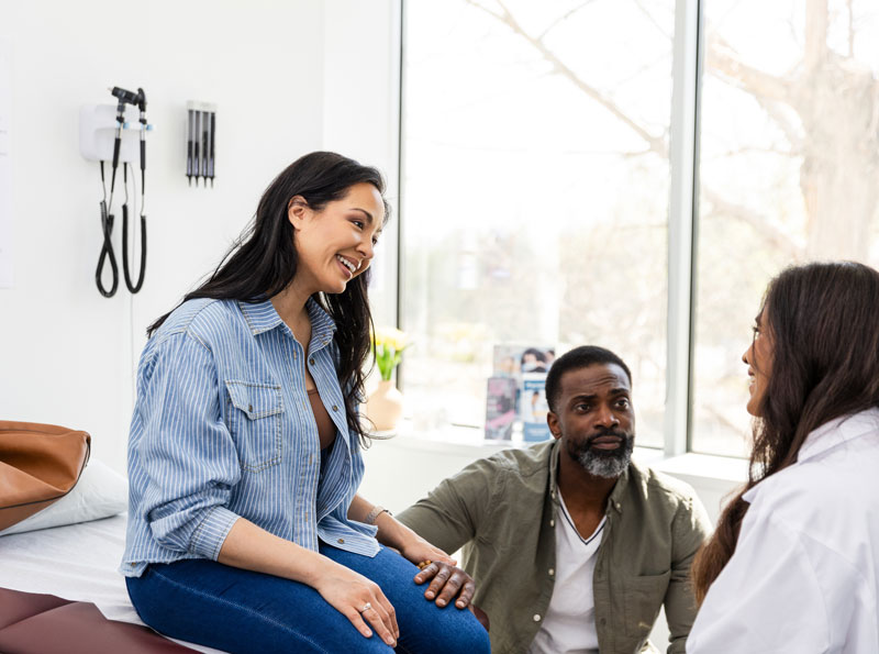 Diverse-couple-sees-female-doctor-about-wife's-symptoms Couple speaking to female doctor in an examination room about fertility testing